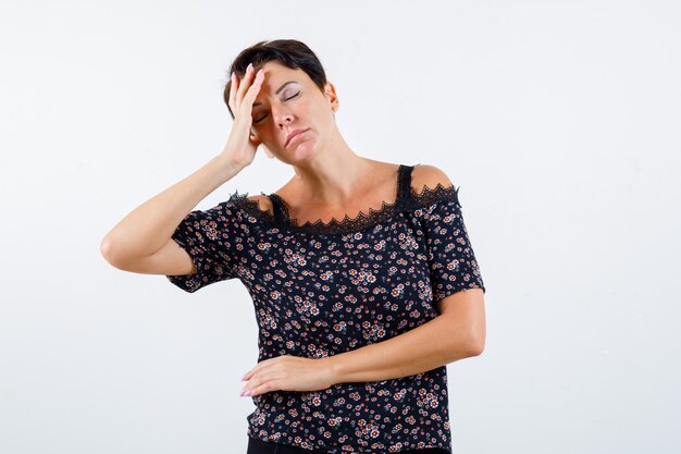 Mujer madura en blusa floral, falda negra con dolor de cabeza y mirando exhausto, vista frontal.