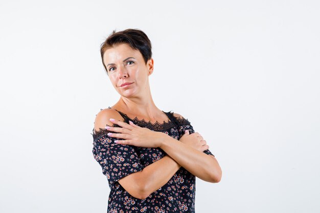 Mujer madura en blusa floral, falda negra sosteniendo dos brazos cruzados sobre el pecho y mirando encantador, vista frontal.