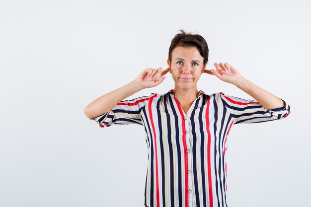 Mujer madura en blusa rayada tapando las orejas con los dedos índices y mirando molesto, vista frontal.