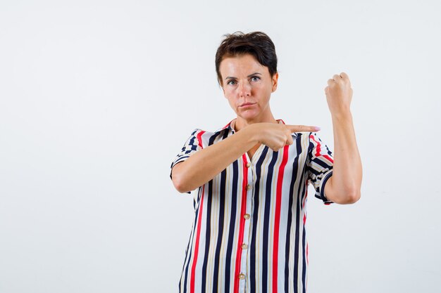 Mujer madura en blusa de rayas apretando el puño, señalando con el dedo índice y mirando confiado, vista frontal.