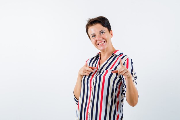 Mujer madura en blusa de rayas apuntando a la cámara con los dedos índices y mirando optimista, vista frontal.