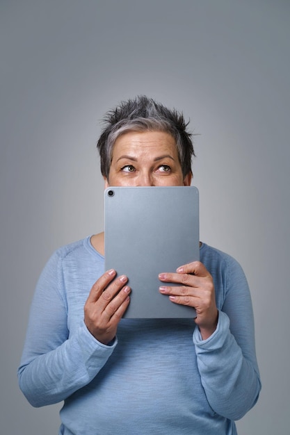 Mujer madura de cabello gris oculta la cara sonriendo tímidamente detrás de un teléfono inteligente o un dispositivo moderno trabajando o comprando en línea o revisando las redes sociales Mujer bonita en blusa azul aislada sobre fondo amarillo