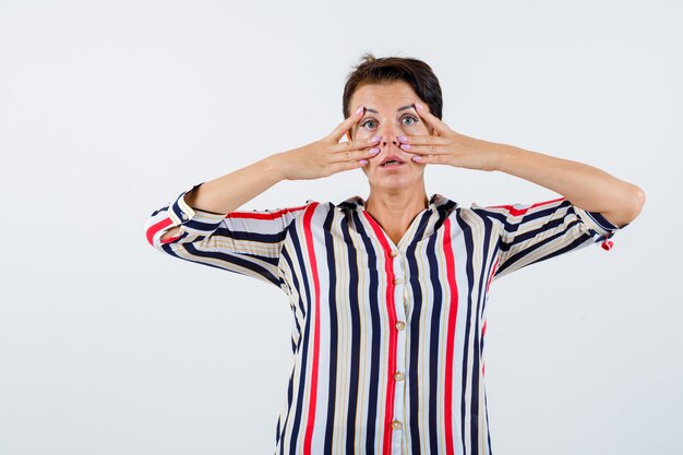 Mujer madura en camisa a rayas mostrando signos v cerca de los ojos y mirando seriamente, vista frontal.