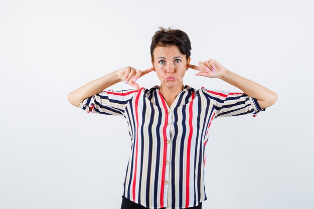 Mujer madura en camisa a rayas taponando los oídos con los dedos índices y mirando serio, vista frontal.