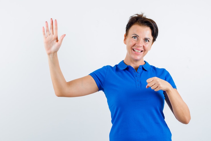 Foto gratuito mujer madura en camiseta azul agitando la mano mientras mantiene el puño cerrado y mirando alegre, vista frontal.
