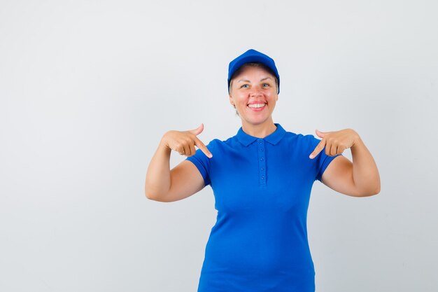 Mujer madura en camiseta azul apuntando hacia abajo y mirando alegre.