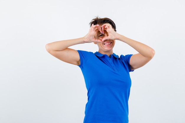 Mujer madura en camiseta azul mostrando gesto de corazón y mirando alegre, vista frontal.