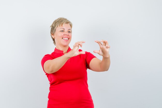 Mujer madura en camiseta roja pretendiendo tomar una foto en el teléfono móvil y mirando alegre.