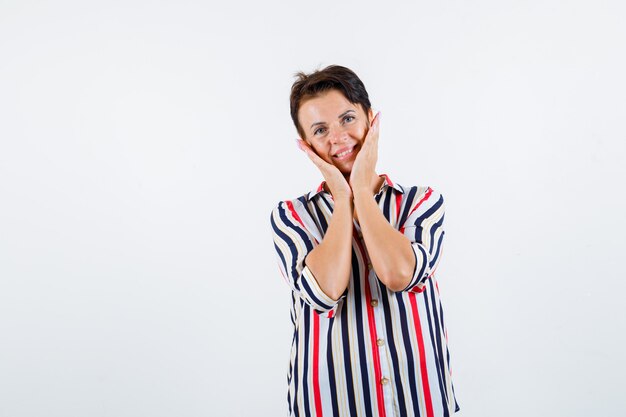 Mujer madura cogidos de la mano en las mejillas en blusa rayada y luciendo optimista, vista frontal.