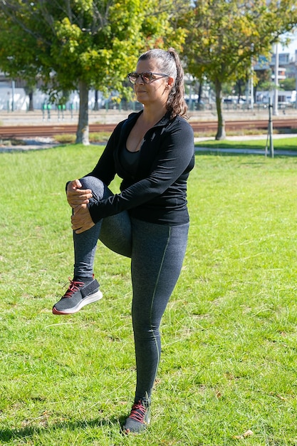 Mujer madura concentrada en ropa de fitness haciendo ejercicio por la mañana en el césped del parque, de pie y estirando las piernas. Concepto de bienestar o estilo de vida activo