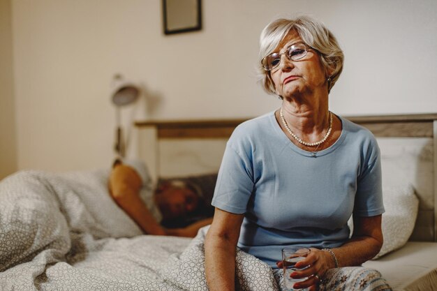 Mujer madura disgustada pensando en algo mientras se sienta en la cama y sostiene un vaso de agua Su marido está durmiendo en el fondo