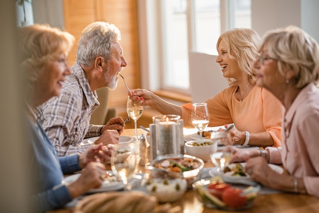 Mujer madura feliz alimentando a su esposo en la mesa del comedor mientras almuerza con sus amigos