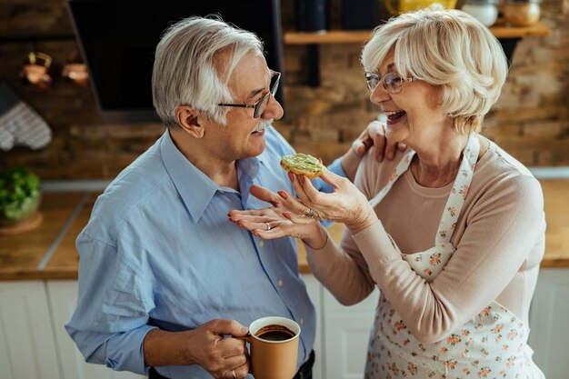 Mujer madura feliz divirtiéndose mientras se comunica con su esposo y lo alimenta en la cocina