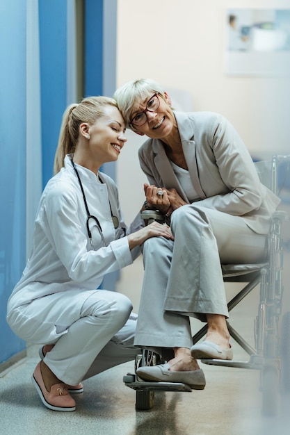 Foto gratuita mujer madura feliz en silla de ruedas cogida de la mano con su doctora en el pasillo del hospital y mirando a la cámara