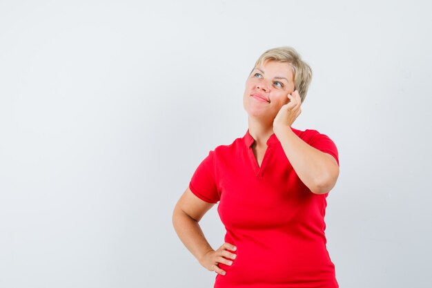 Mujer madura fingiendo hablar por teléfono móvil en camiseta roja y mirando pensativo
