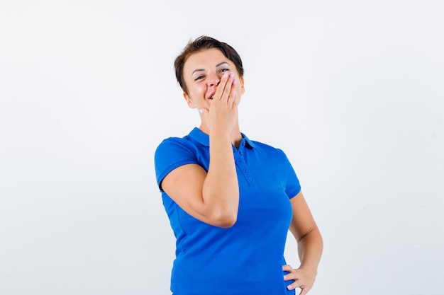 Mujer madura con la mano en la boca en camiseta azul y mirando feliz, vista frontal.