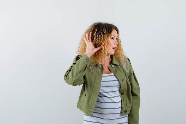 Mujer madura manteniendo la mano detrás de la oreja para escuchar, abriendo la boca con chaqueta verde, camiseta y mirando sorprendido. vista frontal.