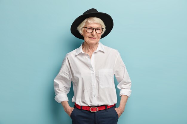La mujer madura de moda viste un elegante sombrero negro, camisa blanca y pantalones con cinturón rojo, mantiene las manos en los bolsillos, tiene una expresión alegre, aislada sobre una pared azul. Concepto de personas, edad y moda.