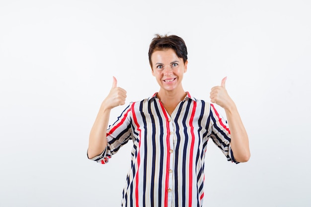 Mujer madura mostrando doble pulgar hacia arriba en camisa a rayas y mirando feliz. vista frontal.