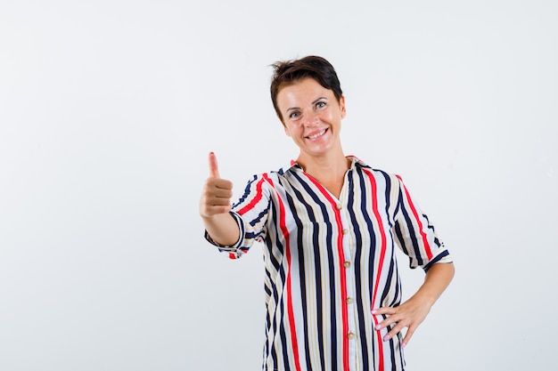 Mujer madura mostrando el pulgar hacia arriba, sosteniendo la mano en la cintura en una blusa a rayas y luciendo optimista, vista frontal.