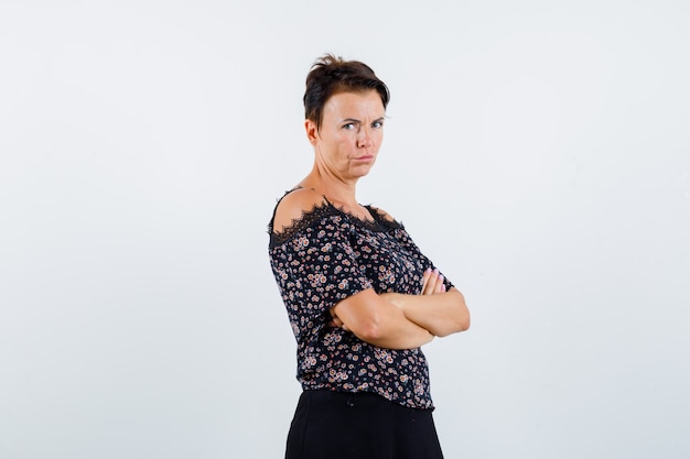 Mujer madura de pie con los brazos cruzados en blusa floral y falda negra y mirando confiado, vista frontal.