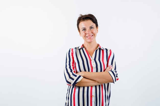 Mujer madura de pie con los brazos cruzados en una blusa a rayas y luciendo optimista, vista frontal.