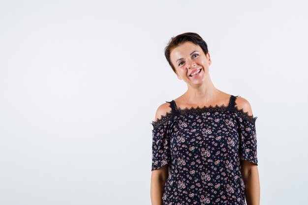 Mujer madura de pie con la espalda recta, sonriendo con blusa floral, falda negra y mirando alegre, vista frontal.