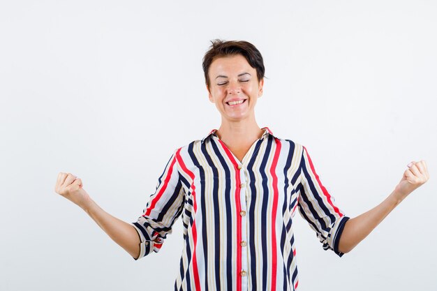 Mujer madura de pie en pose de meditación, cerrando los ojos con una blusa a rayas y mirando alegre. vista frontal.