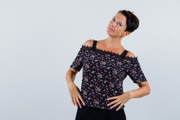 Mujer madura posando manteniendo las manos en la cintura en la blusa y mirando pensativo, vista frontal.