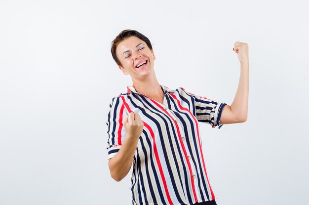 Mujer madura que muestra el gesto del ganador en la blusa rayada y parece afortunado, vista frontal