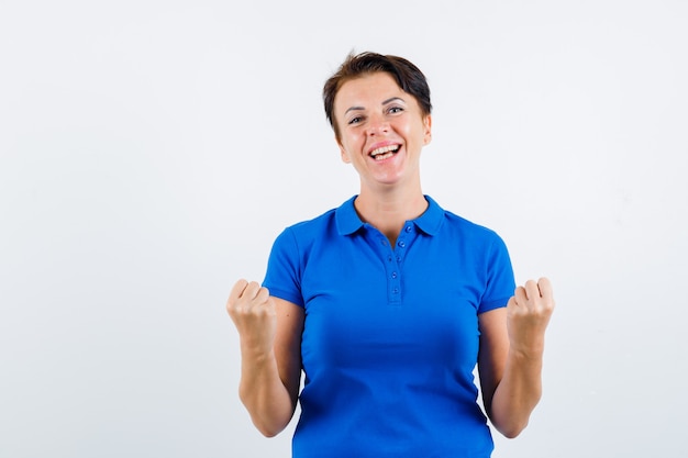 Mujer madura que muestra el gesto del ganador en camiseta azul y parece afortunado. vista frontal.