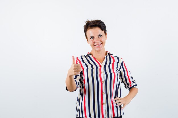 Mujer madura que muestra el pulgar hacia arriba, sosteniendo la mano en la cintura con una blusa a rayas y luciendo confiada. vista frontal.