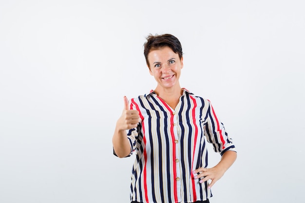 Mujer madura que muestra el pulgar hacia arriba, sosteniendo la mano en la cintura con una blusa a rayas y luciendo confiada. vista frontal.