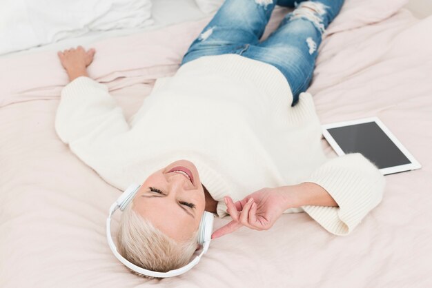 Mujer madura sonriente en la cama disfrutando de escuchar música en auriculares