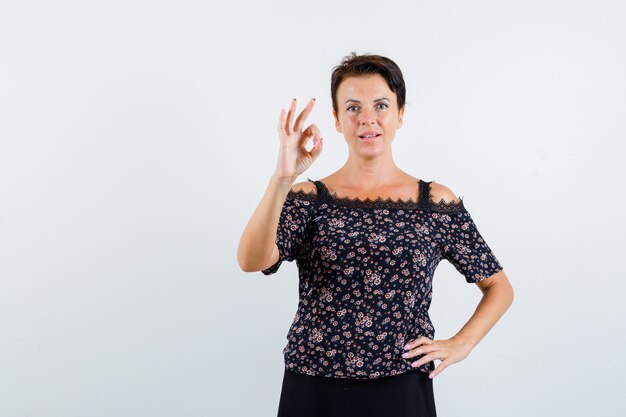 Mujer madura sosteniendo una mano en la cintura, mostrando el signo de ok en blusa floral y falda negra y luciendo confiada. vista frontal.