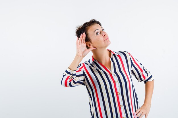 Mujer madura sosteniendo la mano en la cintura, sosteniendo la mano cerca de la oreja para escuchar en blusa rayada y mirando enfocado, vista frontal.