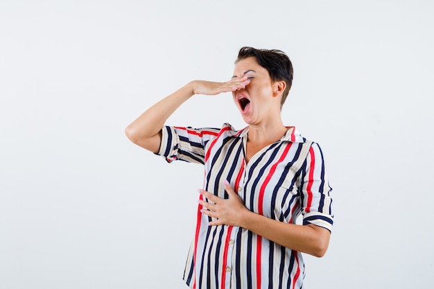 Mujer madura sosteniendo la mano por encima de la boca, tratando de estornudar en blusa rayada y mirando exhausto, vista frontal.