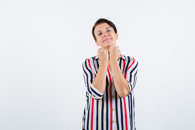 Mujer madura sosteniendo los puños cerrados debajo de la barbilla, sonriendo con blusa a rayas y con un aspecto encantador. vista frontal.