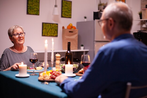 Mujer madura tener una conversación con su marido mientras sostiene una copa de vino tinto en la cocina. Las parejas ancianas sentadas a la mesa en la cocina, hablando, disfrutando de la comida, celebrando su aniversario en