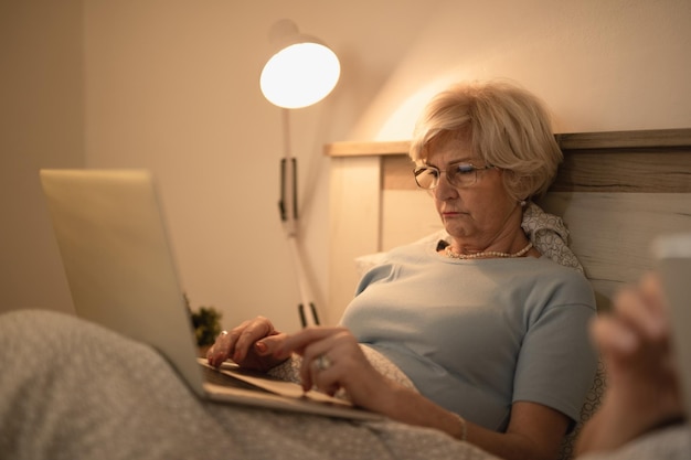 Mujer madura usando una laptop mientras se acuesta en la cama por la noche