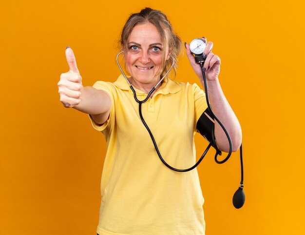 Mujer malsana en camisa amarilla que se siente mejor midiendo su presión arterial con tonómetro sonriendo mostrando los pulgares para arriba sobre la pared naranja