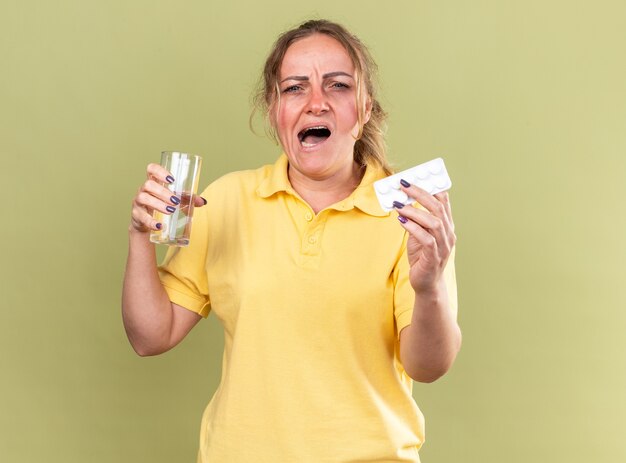 Mujer malsana en camisa amarilla que se siente terrible sosteniendo un vaso de agua y pastillas que sufren de gripe y resfriado de pie sobre la pared verde