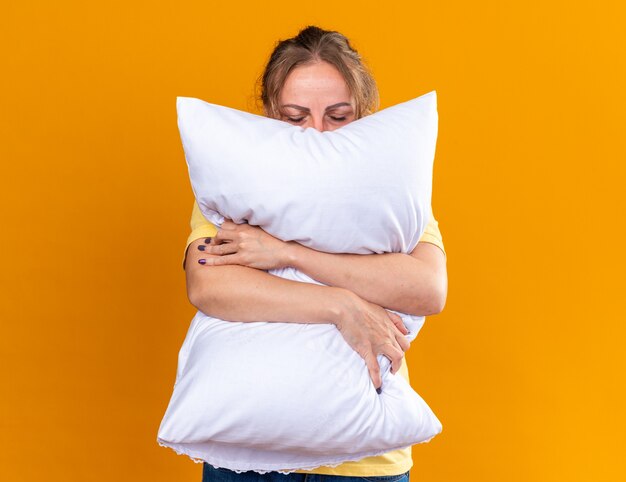 Mujer malsana en camisa amarilla que sufre de gripe y sensación de frío enfermo abrazando la almohada quiere dormir de pie sobre la pared naranja