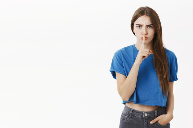 Mujer mandona joven disgustada intensa de aspecto serio con cabello largo y castaño en camiseta azul de moda recortada callando haciendo gesto de shh con el dedo índice sobre la boca pidiendo mantener en secreto