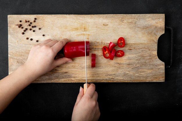 Mujer mano cortar pimiento en tabla de cortar con especias de pimienta en mesa negra