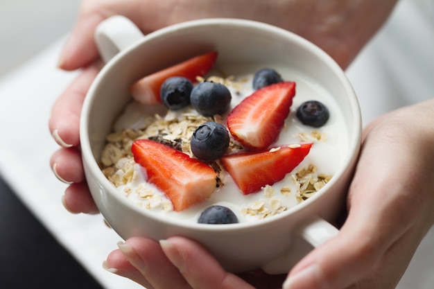 Mujer manos sosteniendo el tazón de fuente con muesli sabroso con las frutas, la avena y el yogur. De cerca. Concepto De Alimentos Saludables.