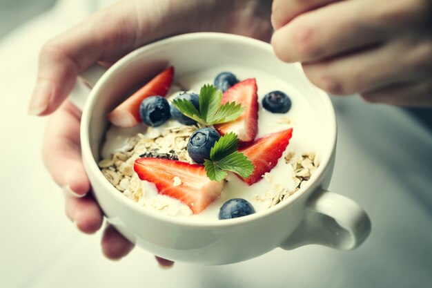 Mujer manos sosteniendo el tazón de fuente con muesli sabroso con las frutas, la avena y el yogur. De cerca. Concepto De Alimentos Saludables.