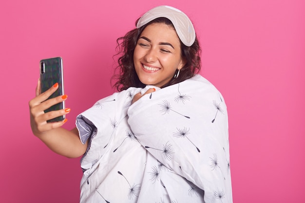 Mujer con manta, adorable mujer con sonrisa encantadora haciendo selfie a través de su moderno teléfono inteligente, mirando su dispositivo