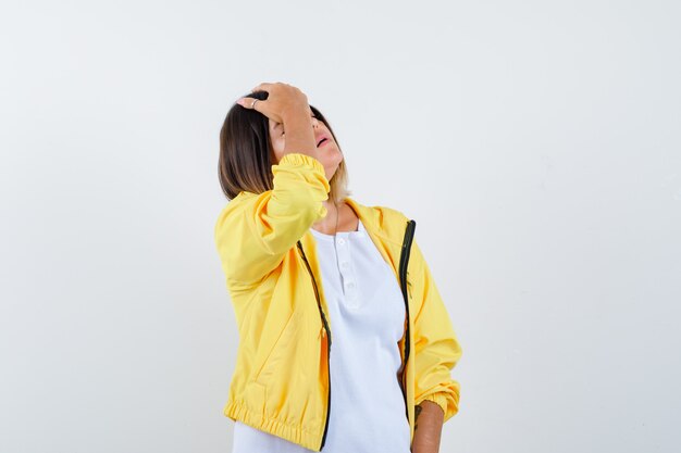 Mujer manteniendo la mano en la cabeza en camiseta, chaqueta y luciendo olvidadiza. vista frontal.