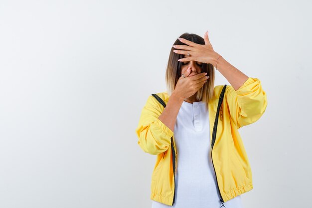 Mujer manteniendo las manos en la boca y la frente en camiseta, chaqueta y luciendo angustiada, vista frontal.